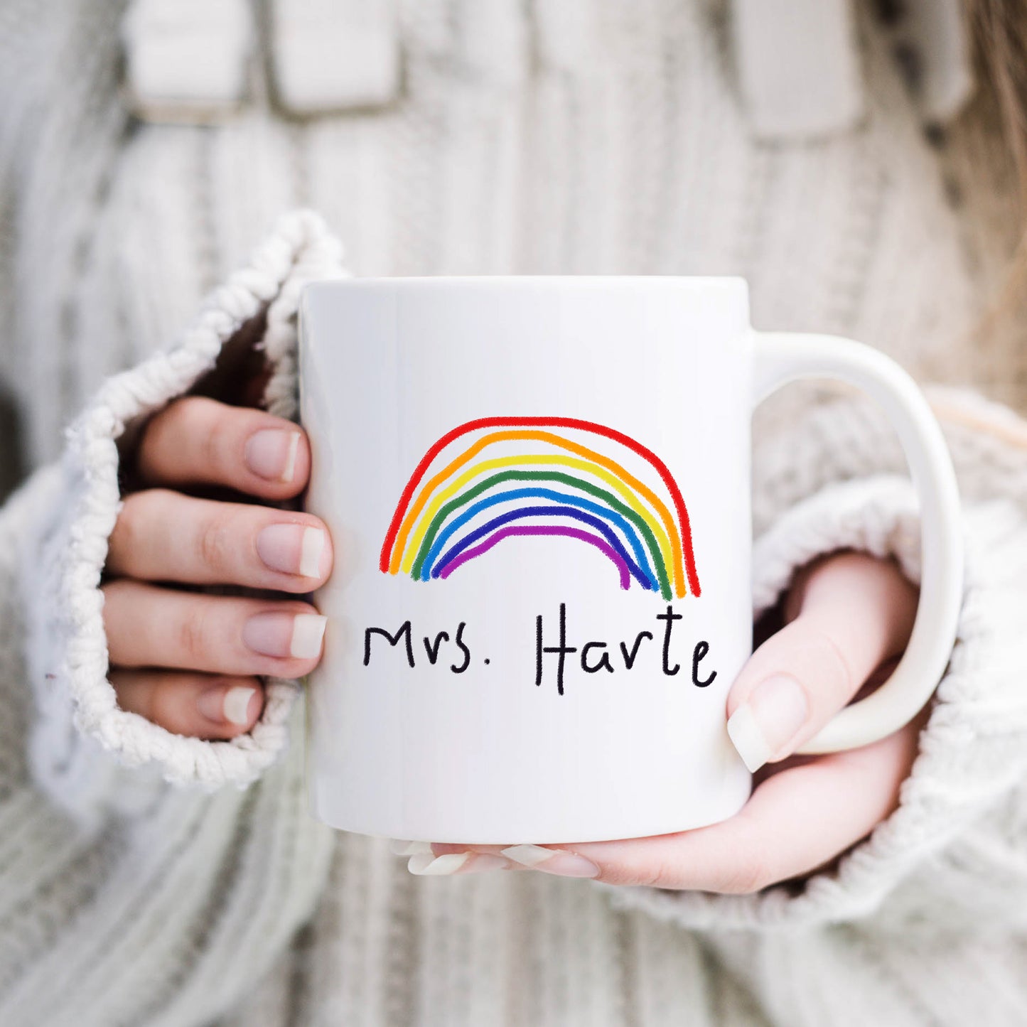 White mug with rainbow design and 'Mrs. Harte' text held by a person in a cozy setting. | Little Heirloom