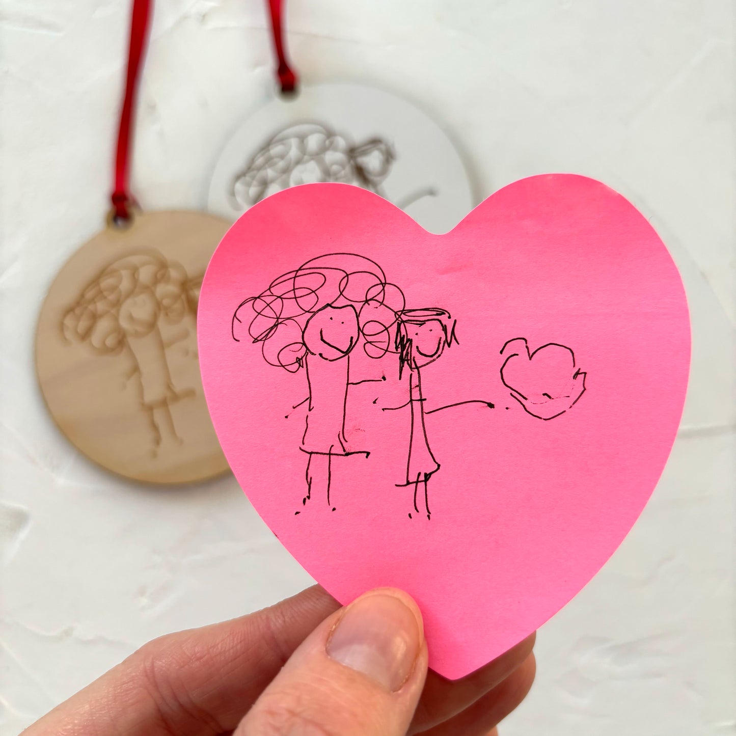 Hand holding a pink heart-shaped card with stick figure drawings of a couple and a heart on a light background. | Little Heirloom