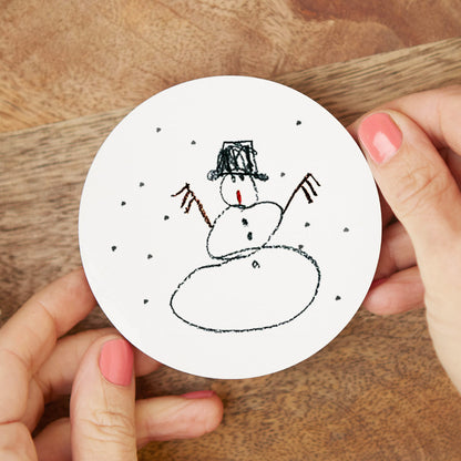 Hand holding a round white coaster with a snowman drawing on a wooden surface | Little Heirloom