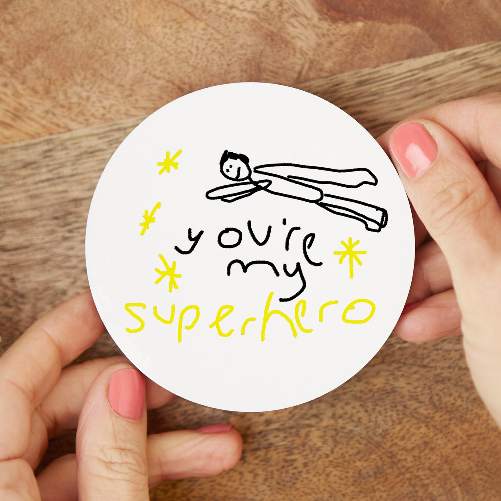 Circular sticker with 'You're my superhero' text and illustration held by hands on a wooden surface| Little Heirloom